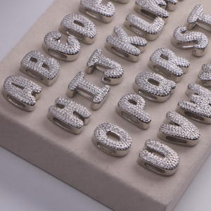 May include: Silver-toned alphabet charms, each encrusted with small, sparkling crystals. The letters are displayed on a neutral-colored surface. The charms are designed with a rounded, block-letter style, perfect for jewelry making or personalized crafts.