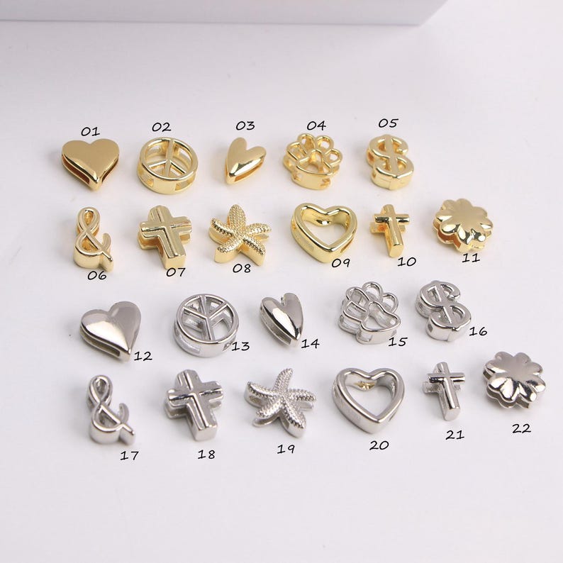 Puede incluir: Una colecci&oacute;n de peque&ntilde;os dijes decorativos en tonos dorados y plateados. Los dijes presentan varias formas, incluyendo corazones, cruces, s&iacute;mbolos de la paz, estrellas de mar y s&iacute;mbolos de d&oacute;lar. Cada dije est&aacute; numerado.