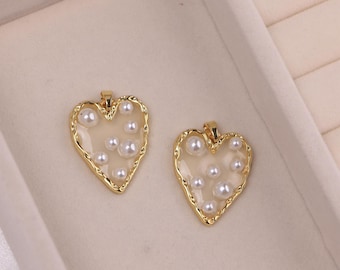 May include: Two heart-shaped pendants with a gold-colored border and filled with small, white pearls. The pendants have a clear, slightly off-white center. The gold border has a textured, irregular edge. The pendants are displayed on a light-colored surface.