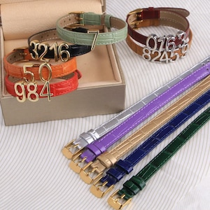 May include: A collection of leather bracelets in various colors, including green, black, orange, and red, with gold-colored numbers attached. Additional bracelets in silver, purple, gold, blue, and green are also displayed, showcasing a variety of styles and designs.