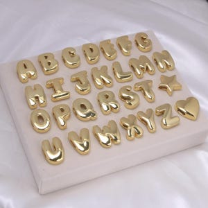 Puede incluir: Una colección de abalorios dorados con letras del alfabeto, incluyendo una estrella y un corazón. Las letras están en una fuente redondeada, tipo burbuja, expuestas sobre una superficie de color claro. Ideal para la creación de joyas o manualidades personalizadas.