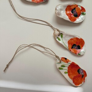 May include: Hand-painted oyster shell ornaments featuring vibrant red poppy flowers with black centers and green leaves. Each shell is strung with natural twine for hanging. Decorative coastal-themed art.
