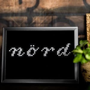 Puede incluir: Una obra de arte enmarcada en negro con la palabra "nörd" bordada en punto de cruz blanco. El marco es gris oscuro y la obra se exhibe sobre un fondo rústico con un jarrón decorativo y un cuadro enmarcado.
