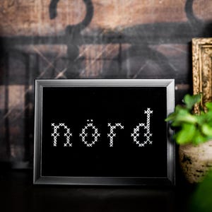 Puede incluir: Una obra de arte en punto de cruz negro enmarcada con la palabra "nörd" en blanco. La obra se exhibe sobre una superficie oscura, con una escultura de mano decorativa y una imagen enmarcada en el fondo.