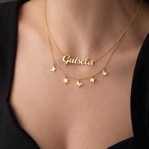 May include: A gold-coloured layered necklace. The top layer has the name "Guisela" in cursive. The bottom layer features small star and butterfly charms. The necklace is worn against a dark background.