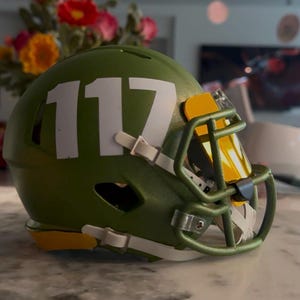 May include: An olive green football helmet featuring the white number 117 printed on the side. The helmet includes a matching olive green face mask, white chin strap clips, and a reflective gold-tinted visor attached to the front of the mask.