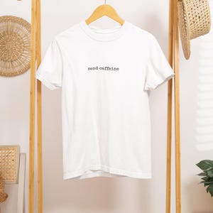 May include: A white t-shirt with the phrase "send caffeine" printed in black. The shirt is hanging on a wooden clothes rack with a straw hat and woven wall decor. Folded clothing is on the rack's shelf.