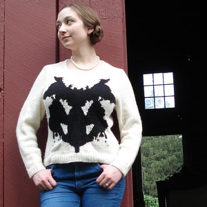 May include: A woman wearing a white sweater with a black abstract design. The sweater has long sleeves and a crew neck. She is wearing blue jeans and has a pearl necklace on.