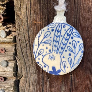Puede incluir: Adorno navideño de cerámica con un diseño pintado a mano. El adorno presenta un diseño de conejo azul y blanco con detalles florales. Cuelga de una cuerda blanca sobre un fondo de madera rústica.