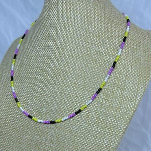 May include: A beaded necklace in the colours of the non-binary pride flag. The necklace is made of small, round beads in black, white, yellow, and purple. The necklace is displayed on a mannequin.