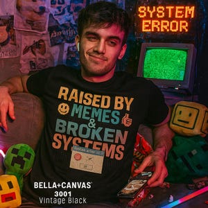 以下が含まれることがあります： オレンジ、ティール、白で「RAISED BY MEMES & BROKEN SYSTEMS」と書かれた黒いTシャツ。シャツには「BELLA+CANVAS 3001 Vintage Black」の文字も入っています。その人は携帯電話を持っています。