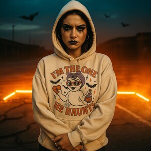 May include: A beige hoodie with the text "I'M THE ONE HE HAUNTS" and a cartoon graphic of a baby wearing sunglasses, holding a heart and a potion bottle. The hoodie has a hood and is worn by a person.