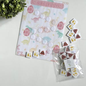 Peut inclure: Un tableau de propreté sur le thème des dinosaures. Le tableau comporte une bannière rose avec l'inscription "Je suis propre !" et comprend des cercles velcro pour fixer des autocollants de récompense. Un petit sac contient des autocollants avec des images de caca et d'urine.