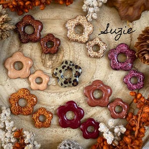 May include: A collection of flower-shaped resin pieces in various colors and finishes, including glitter, solid, and patterned designs. The pieces are arranged on a wooden surface with dried floral accents. The name "Suzie" is written in cursive.