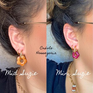 May include: Two close-up shots of earrings. The left earring is a gold hoop with an orange flower charm, labeled "Mid Suzie." The right earring is a gold hoop with a pink flower charm, labeled "Mini Suzie." The text "Cr&eacute;ole Hossegoria" is at the top.