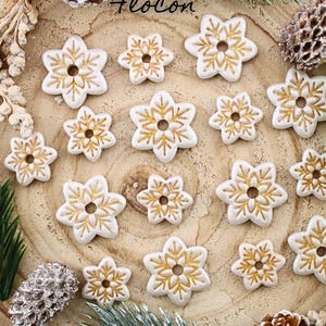 May include: White snowflake-shaped ornaments with gold detailing are arranged on a wooden surface. The ornaments vary in size and are surrounded by pine needles, pine cones, and the word "Flocon".