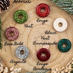 May include: A selection of donut-shaped decorations in shades of green, red, rose gold, silver, and burgundy, displayed on a wooden base. The word "Donut" is written at the top. Colour names are written in French.