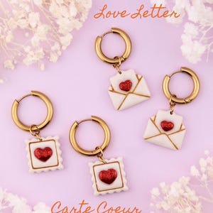 Peut inclure: Boucles d'oreilles créoles dorées avec des breloques en forme d'enveloppe blanches et dorées, ornées de cœurs rouges pailletés. Le fond est rose, avec les mots "Love Letter" et "Carte Coeur".