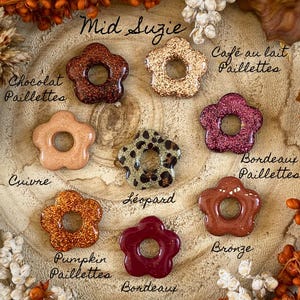 May include: A collection of flower-shaped resin beads in various colors and finishes, including glitter and leopard print. The beads are arranged on a wooden surface and labeled with names like "Mid Suzie", "Pumpkin Paillettes", and "Bordeaux".