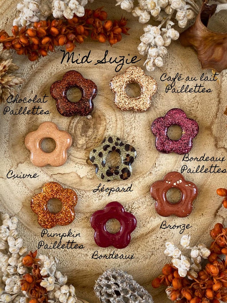May include: A collection of flower-shaped resin beads in various colors and finishes, including glitter and leopard print. The beads are arranged on a wooden surface and labeled with names like "Mid Suzie", "Pumpkin Paillettes", and "Bordeaux".