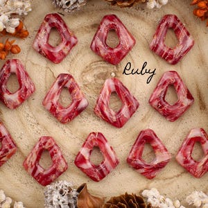 Peut inclure: Des pendentifs en forme de losange rouge et blanc avec un trou central et des paillettes dorées. Le mot "Ruby" est écrit en cursive. Les pendentifs sont disposés sur une surface en bois avec des pommes de pin et des fleurs séchées.