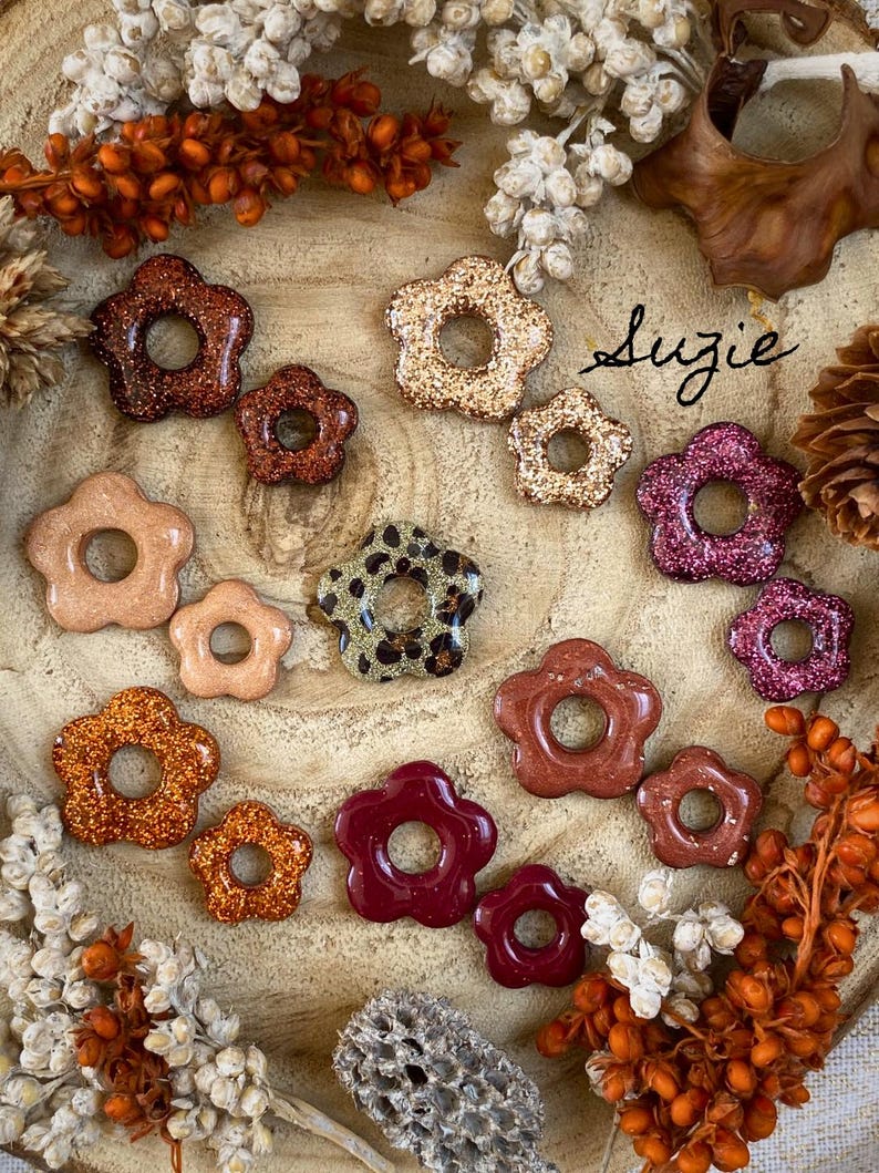 May include: A collection of flower-shaped resin pieces in various colors and finishes, including glitter, solid, and patterned designs. The pieces are arranged on a wooden surface with dried floral accents. The name "Suzie" is written in cursive.