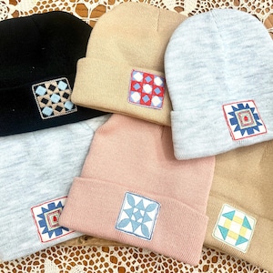 May include: A collection of beanies in various colors, including black, tan, pink, and gray. Each beanie features a square embroidered patch with geometric designs in contrasting colors. The beanies are displayed in a wooden box.