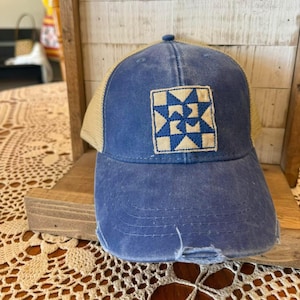 May include: A distressed blue baseball cap with a tan mesh back. The front features a square patch with a blue and tan quilt block design. The cap has a pre-worn look with a distressed brim.