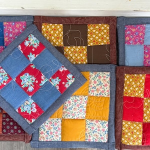 May include: A collection of handmade quilted pot holders. Each square pot holder features a patchwork design with various floral and geometric patterns in red, blue, yellow, and brown. The edges are finished with a denim border.