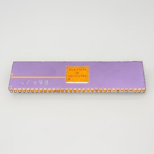 Puede incluir: Un chip de circuito integrado rectangular de color morado con una placa cuadrada central dorada que contiene el texto USA 8543A, SBP9989NJ y un pequeño símbolo. El número 47998 está impreso a la izquierda y una fila de pines dorados bordea la base.