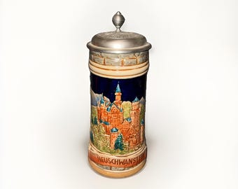 May include: A ceramic beer stein featuring a relief depiction of Neuschwanstein Castle against a dark blue background. The vessel has a metallic lid with a finial and a base with a stone-like texture. The text NEUSCHWANSTEIN is printed at the bottom.