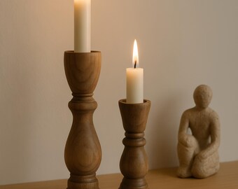 May include: Two wooden candle holders with lit white candles. The candle holders are different sizes and have a turned wood design. A small sculpture of a person is on the shelf in the background.