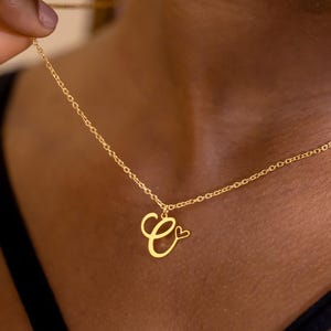 Può includere: Una collana color oro con un ciondolo a forma di lettera "C" corsiva e un piccolo cuore. La delicata catena è tenuta da una mano, mettendo in risalto il design del ciondolo. La collana è un accessorio semplice ed elegante.