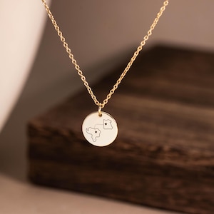 Puede incluir: Un collar de tono dorado con una cadena fina y un colgante circular. El colgante está grabado con dos contornos de mapas conectados por una línea discontinua, cada uno con un pequeño símbolo de corazón centrado dentro de las formas terrestres.