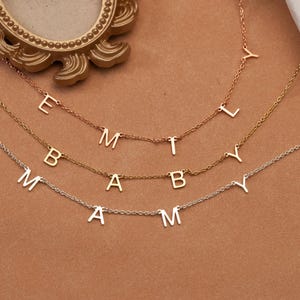 Puede incluir: Tres delicados collares con colgantes de letras individuales que deletrean nombres. Los collares son en tonos dorados, oro rosa y plateado. Los nombres son "Emily", "Baby" y "Amy". Los collares se muestran sobre un fondo neutro.