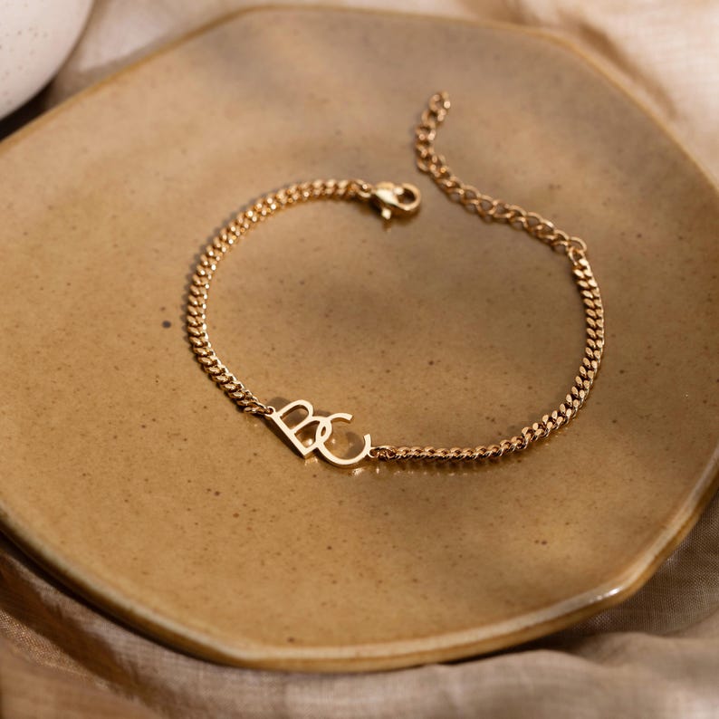 May include: Gold-colored chain bracelet with the letters "BC" in a delicate font. The bracelet is displayed on a brown plate, creating a warm, neutral backdrop. The chain has a classic curb link design, and the clasp is visible.