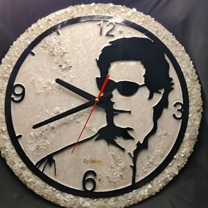 May include: A round wall clock featuring a black silhouette of a person wearing sunglasses. The clock face is light-colored with a textured surface and black numerals. The clock hands are black, with a red second hand. The clock's outer edge is decorated with small, clear crystals.