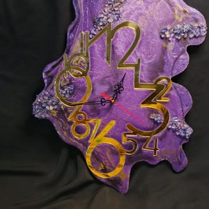 Puede incluir: Reloj de pared de forma abstracta con un diseño de resina morada y dorada. El reloj presenta números y manecillas doradas, con detalles decorativos azules y blancos. Las dimensiones del reloj no son visibles.