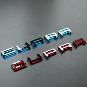 以下が含まれることがあります： 「SUPRA」の文字を綴る、金属製の車のエンブレムのクローズアップ。エンブレムは黒色のベースに、青と赤のアクセントが光っています。文字は灰色の表面に配置されています。