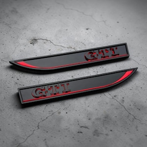 Könnte beinhalten: Zwei längliche, mattschwarze Autoembleme mit den Buchstaben GTI in einer fetten, erhabenen Schrift. Jedes Emblem hat ein spitz zulaufendes Ende und eine leuchtend rote Akzentlinie, die horizontal über die Oberfläche verläuft.