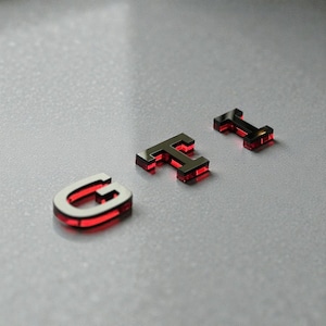 Peut inclure: Gros plan sur les lettres G, T et I, chacune avec une finition argentée métallique et une lueur rouge vibrante. Les lettres sont disposées sur une surface gris clair, créant un design moderne et accrocheur.