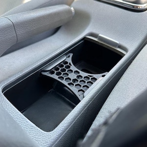 Puede incluir: Un organizador de consola de coche negro con un diseño de panal. El organizador se coloca dentro de la consola central de un coche, rodeado de plástico y tela grises. El patrón de panal es visible en la parte superior del organizador.