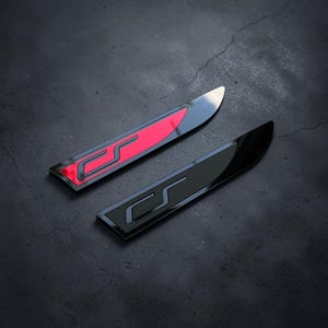 Pode incluir: Dois emblemas alongados e afilados com um design de letra S estilizada. Um emblema tem um acabamento preto brilhante com uma incrustação vermelha vibrante, enquanto o segundo emblema é totalmente acabado em material preto brilhante refletor.