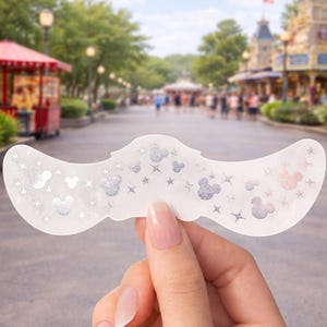 May include: A white eye mask with a curved shape, adorned with silver Mickey Mouse designs and stars. The mask is held up against a blurred backdrop of a theme park, with trees and buildings visible.