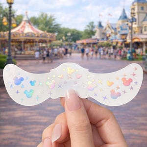 May include: A white, iridescent sticker with a curved shape, featuring Mickey Mouse silhouettes and stars in pastel colors. The sticker is held up against a blurred background of a theme park.
