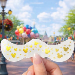 May include: A white decorative item with gold Mickey Mouse silhouettes and star accents. The item is held up against a blurred background of a park with balloons and buildings, suggesting a theme park souvenir.