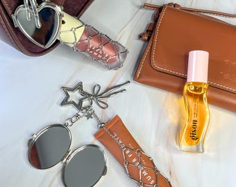May include: An assortment of beauty products and accessories, including lip gloss, perfume, and a small mirror. The lip gloss is encased in a silver net, and the perfume bottle has a pink cap. The items are arranged on a marble surface.