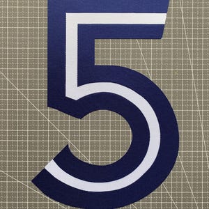 Peut inclure: Un grand écusson en tissu bleu marine en forme de chiffre cinq, doté d'un contour blanc audacieux qui suit la forme du chiffre. L'écusson présente une texture uniforme et nette, présenté sur une surface à motif quadrillé pour référence.