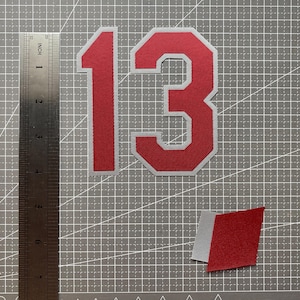 May include: A red and silver number 13 patch with a block font style, accompanied by a small red and silver diamond-shaped patch. A metal ruler is positioned to the left, showing the number 13 patch is approximately 15.2 cm in height.