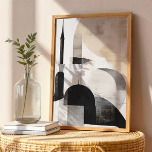 May include: Abstract art print in a wooden frame. The artwork features black, white, and beige geometric shapes. A clear glass vase with greenery and a stack of books are in the foreground, placed on a woven table.
